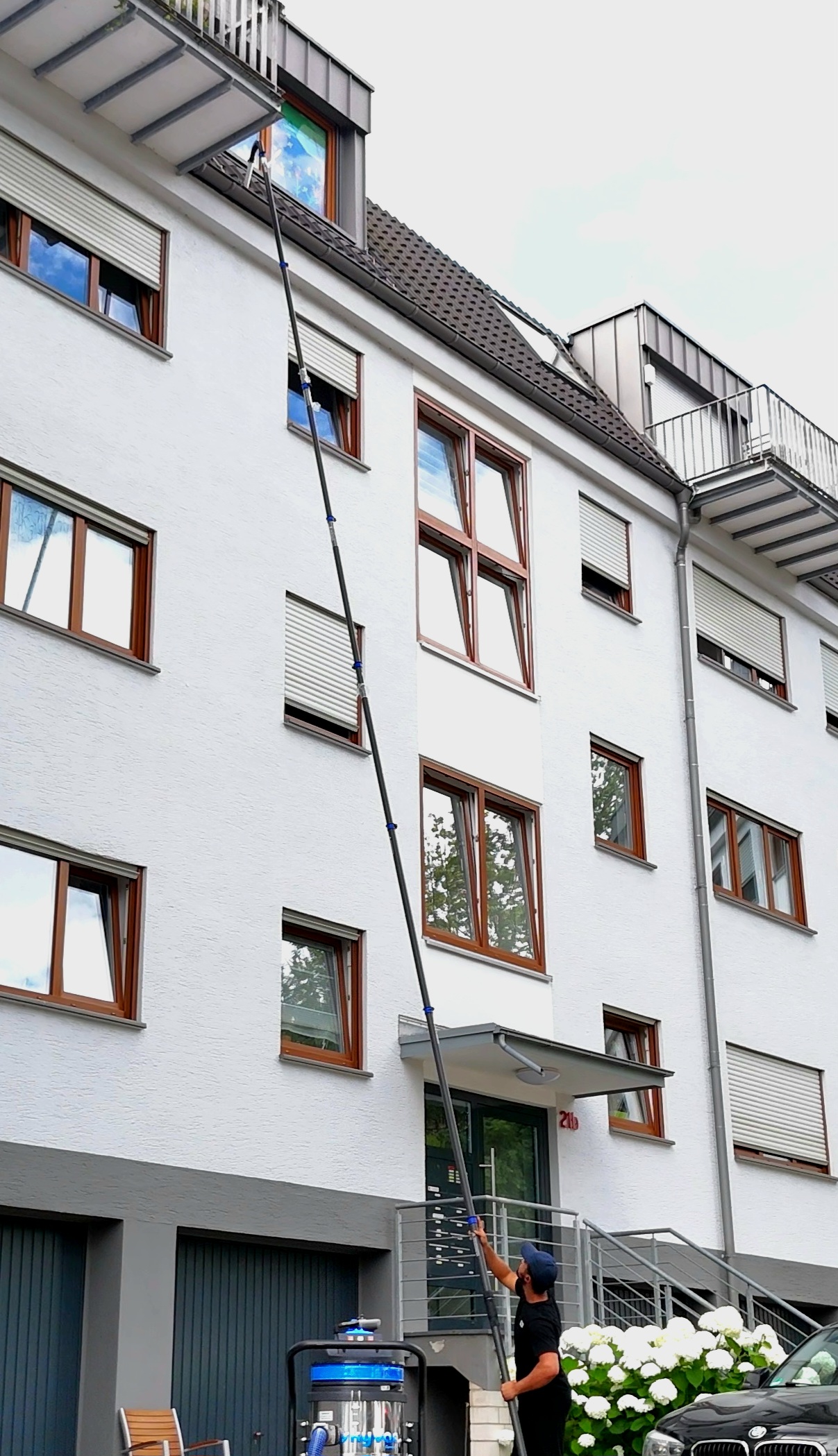 Wide-angle, photographic shot of a detached family home in a quiet German residential street typical for the Siegen region, with rolling green hills softly blurred in the background. The camera is placed at ground level, looking slightly upward along the line of the roof so the complete gutter run is visible. In the foreground stands a tidy, robust gutter vacuum machine with long, segmented poles laid out beside it, ready for use, emphasizing preparation for a quick appointment. Late afternoon natural light casts gentle, warm highlights on the house façade and equipment, creating a calm, reliable atmosphere. The composition follows the rule of thirds, with sharp focus throughout to showcase the property, gutters, and ground-based system in a clean, professional style.